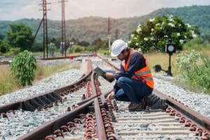 A worker wearing a hard hat and high vis jacket kneels on a train track and touches the railway while holding a clipboard